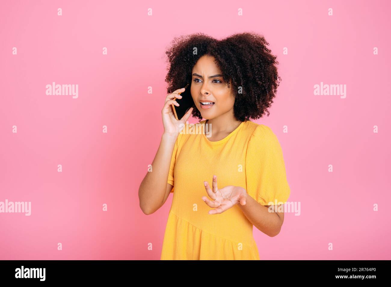 Telefongespräch. Verwirrte, erfolgreiche, fleißige, lockige Frau, die mit einem Freund oder Kollegen am Handy spricht, wegsieht, mit der Hand Gesten macht, auf einem isolierten rosa Hintergrund steht Stockfoto