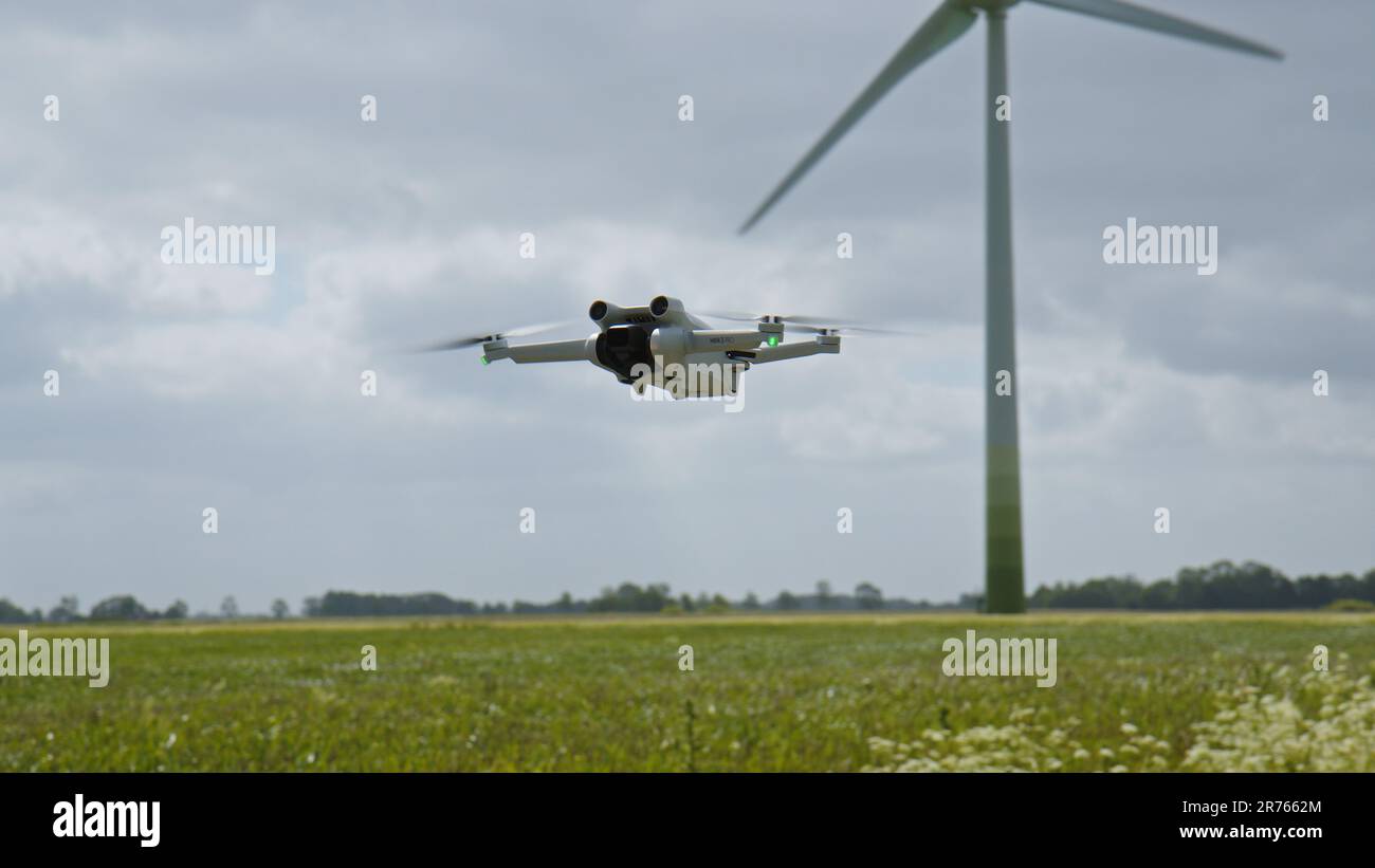 OSTFRIESIEN, DEUTSCHLAND - 25. MAI 2023: Kamera Drohne DJI Mini 3 Pro schwebt in der Luft in der Nähe eines Feldes mit Windturbinen Stockfoto