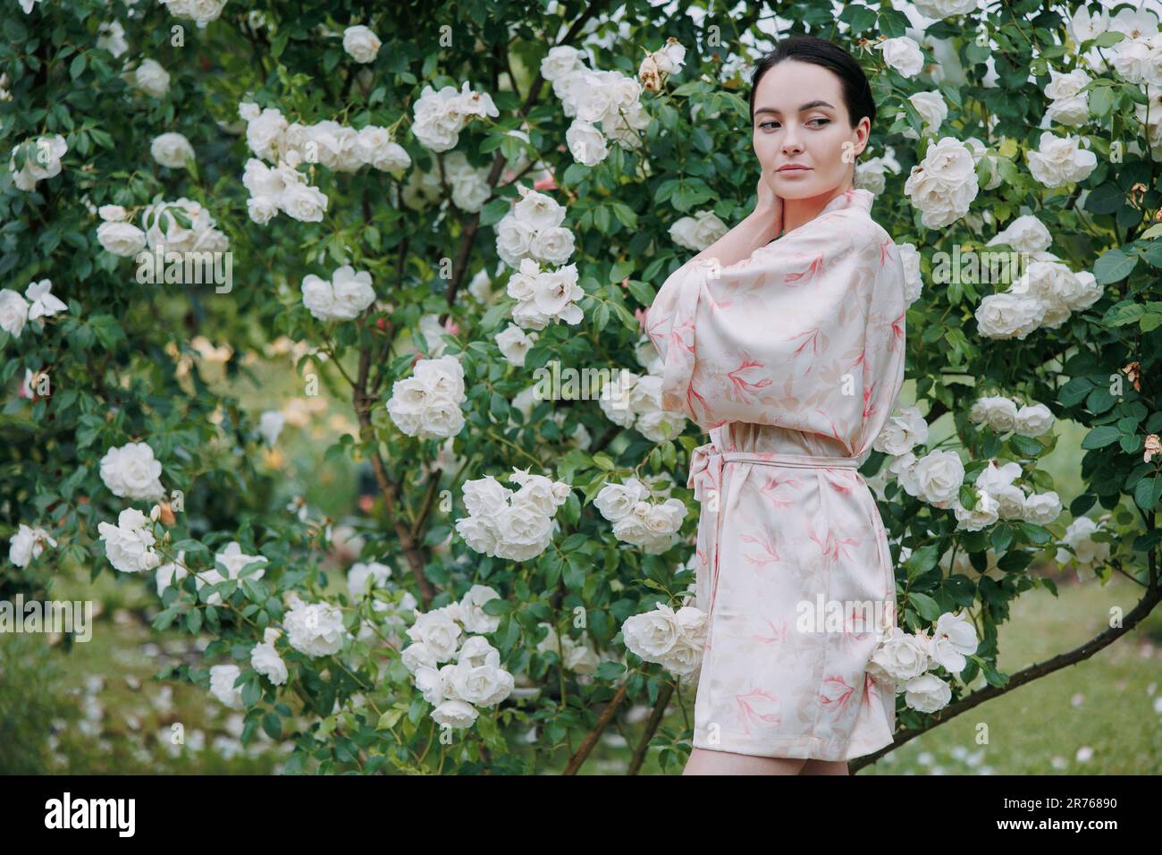 Junge brünette Frau im Kimono-Kleid, die sich an der blühenden, weißen Eisbergrose im Garten erfreuen kann. Stockfoto