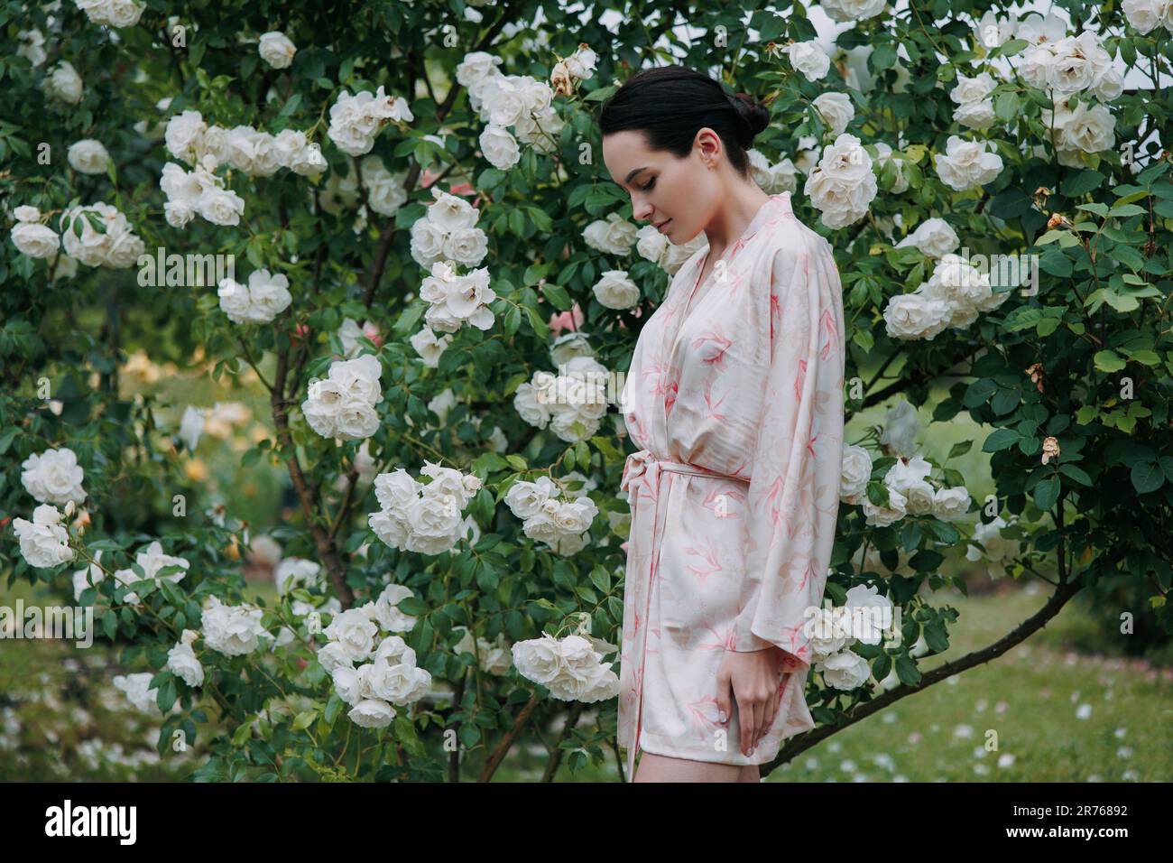 Junge brünette Frau im Kimono-Kleid, die sich an der blühenden, weißen Eisbergrose im Garten erfreuen kann. Stockfoto