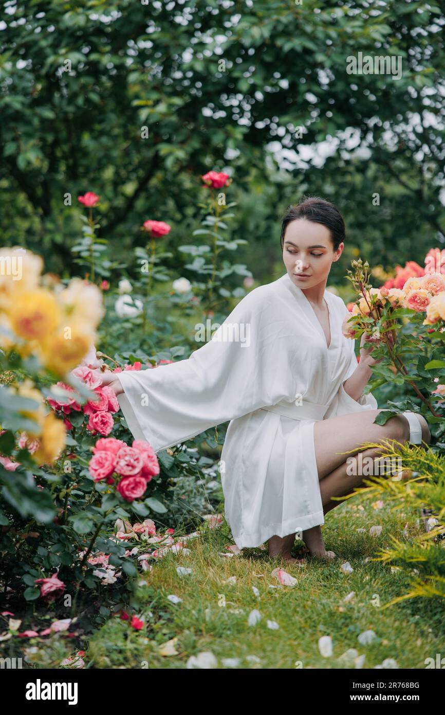 Junge brünette Frau im Kimono-Kleid, die barfuß auf dem Rasen zwischen Rosen im Garten sitzt und die Blüte genießt. Stockfoto