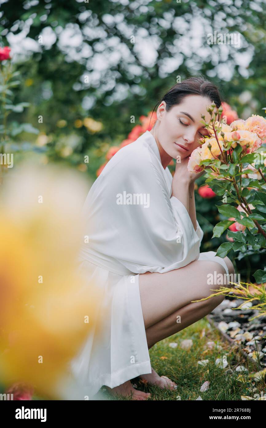 Junge brünette Frau im Kimono-Kleid, die barfuß auf Gras zwischen Rosen im Garten sitzt und die Blüte mit geschlossenen Augen genießt. Stockfoto