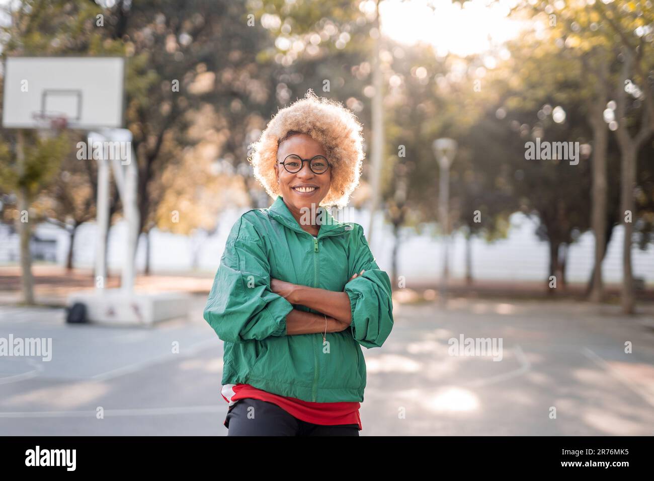 Glückliche junge afroamerikanische Frau in Sportjacke und Brille, die die Arme kreuzt und an sonnigen Tagen im Park in die Kamera schaut Stockfoto