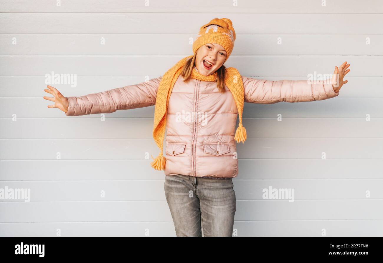 Ausdrucksstarkes Kind, das vor weißem Hintergrund posiert, eine weiche, pinkfarbene, gepolsterte Jacke und einen orangefarbenen Strickmütze und Schal trägt. Wintermode für Kinder Stockfoto