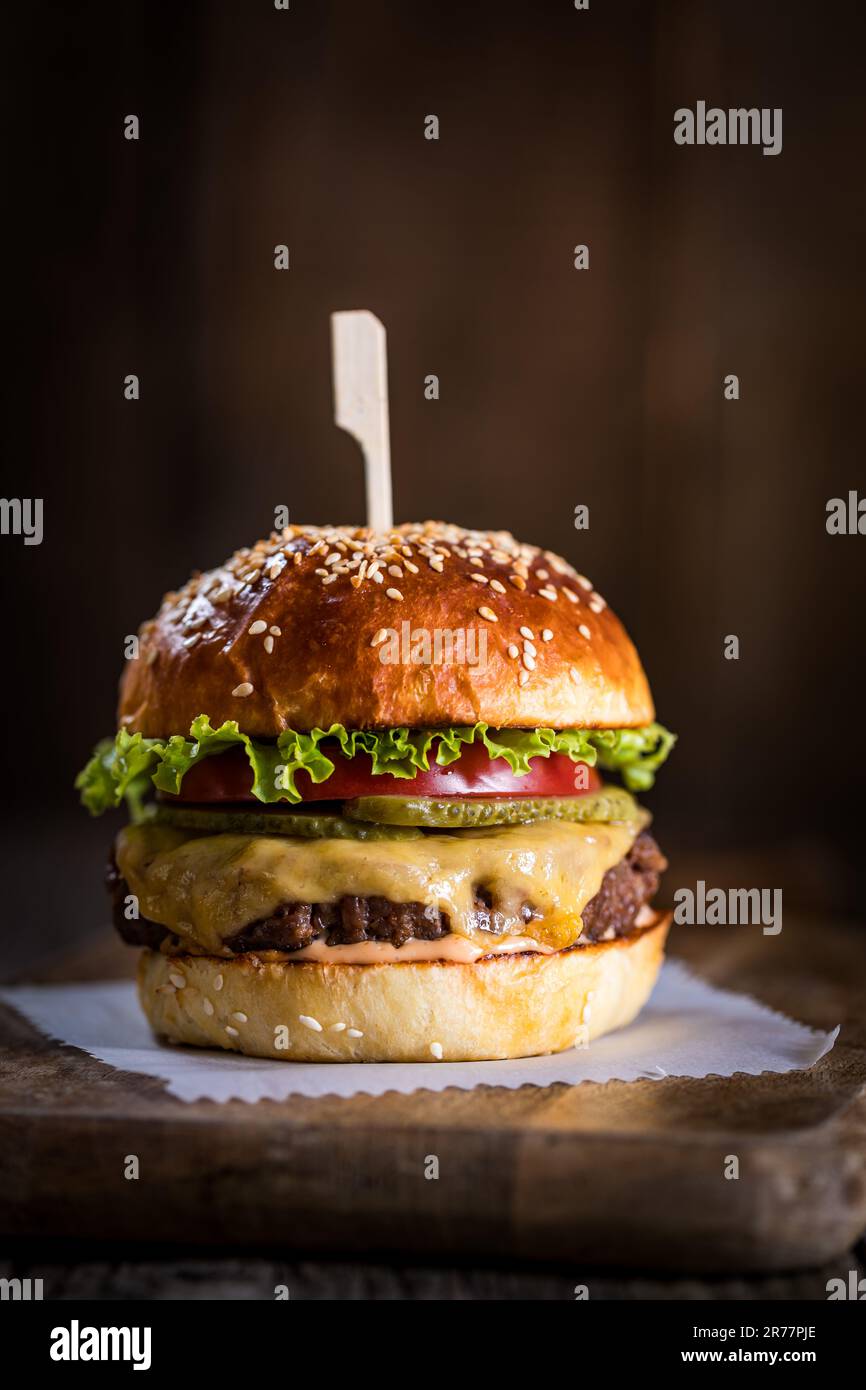 Leckerer Cheeseburger mit Salat, Cheddar, Tomaten und Gurken. Burgerbrötchen mit Sesamsamen. Rustikale Atmosphäre. Köstliches Fast Food Stockfoto