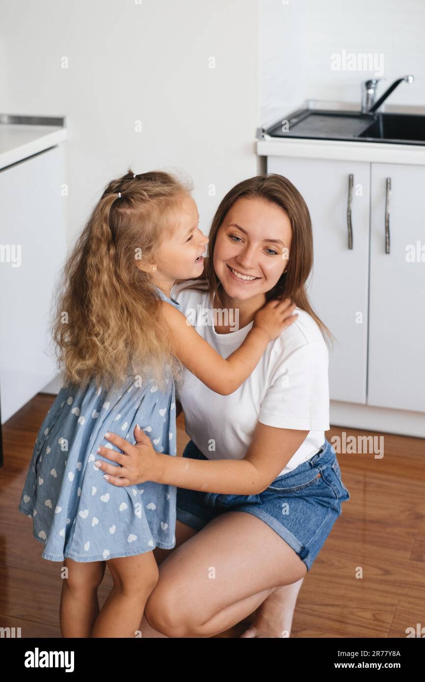 Das Kind flüstert Mutter zu Hause in der Küche etwas ins Ohr. Der Junge erzählt ein Geheimnis. Ein kleines Mädchen, das mit mom spricht und gerne um Erlaubnis bittet Stockfoto