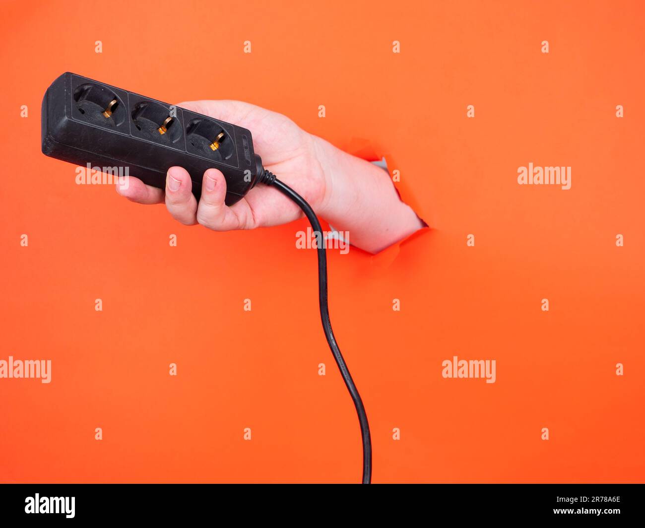 Eine Hand hält eine Steckdosenleiste durch zerrissenes orangefarbenes Papier. Kein Gesicht, Kopierraum. Stockfoto