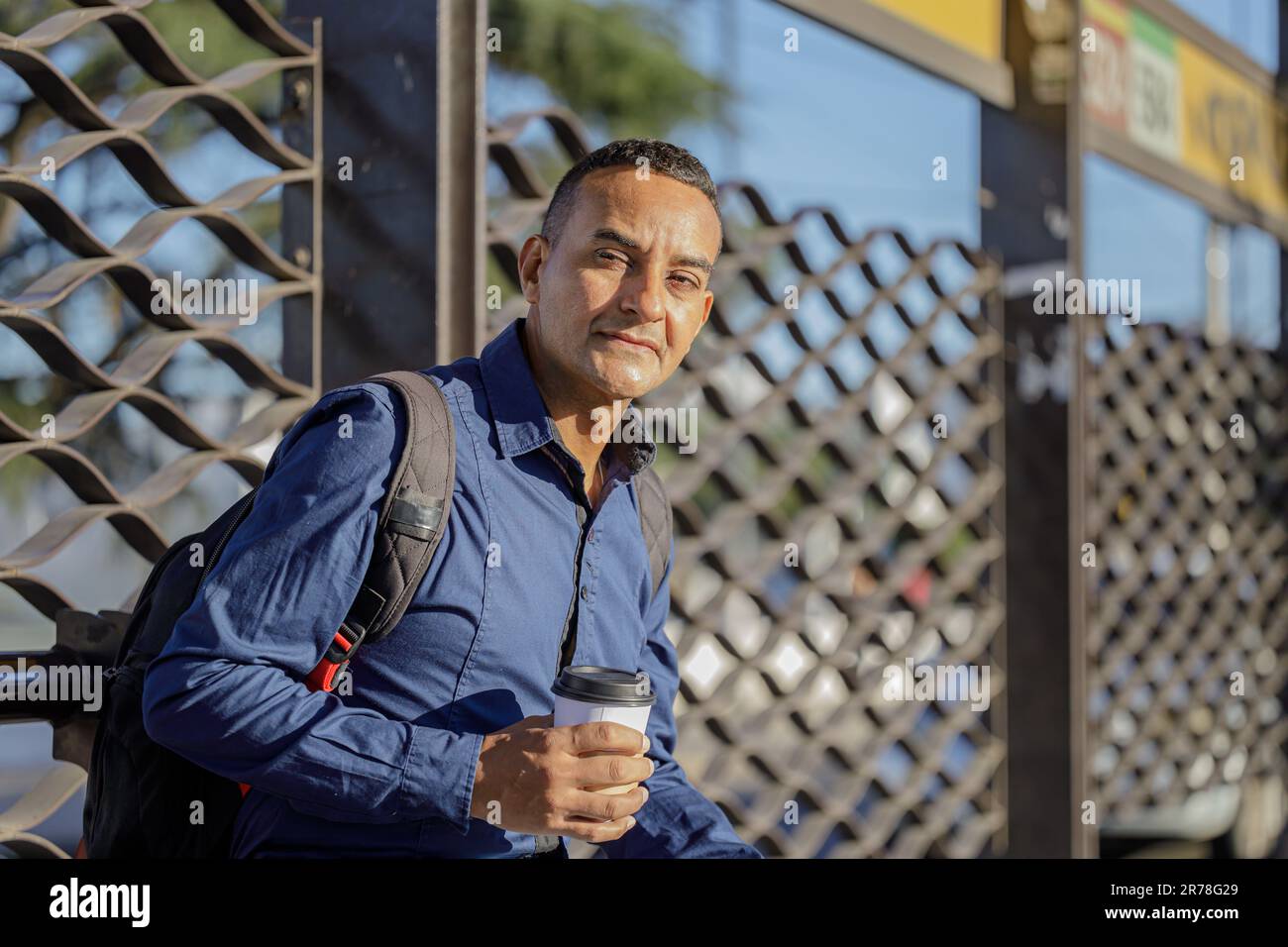 Ein junger latino, der mit einer Papptasse Kaffee in der Hand auf den Bus wartet. Stockfoto