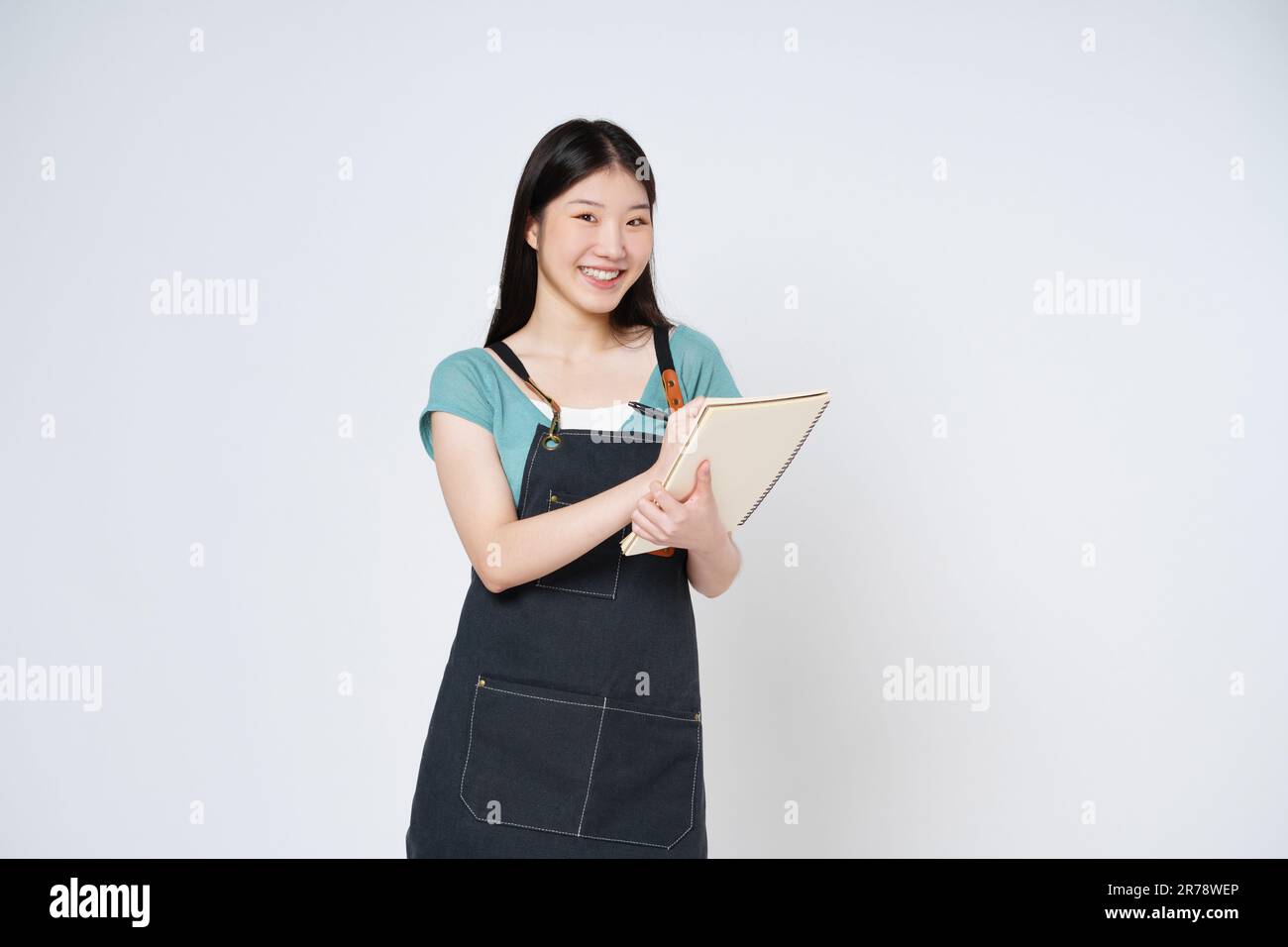 Junge Frau trägt Küchenschürze mit Notizblock, Kochbuch und Stift isoliert auf weißem Hintergrund. Stockfoto
