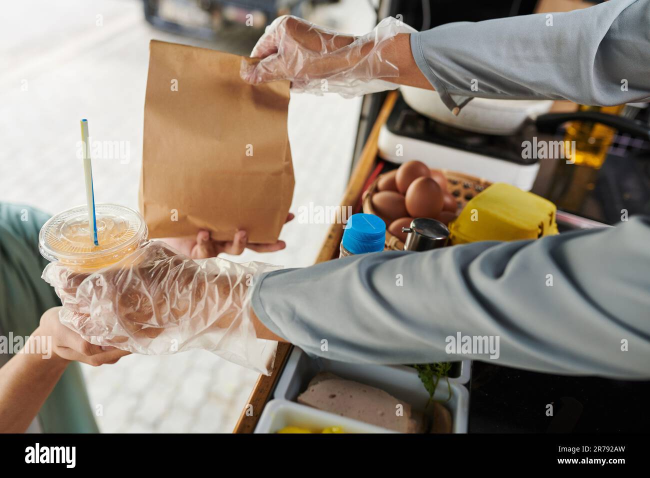 Nahaufnahme der Handschuhe eines Lkw-Verkäufers, der Limonade und Fast Food in einer Papiertüte über Behältern mit Zutaten für Hotdogs verteilt Stockfoto