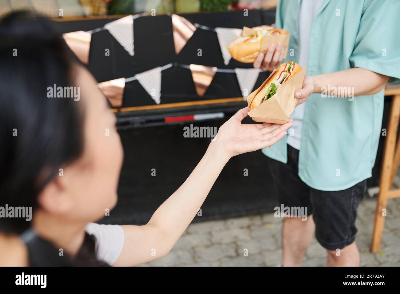Nahaufnahme einer jungen Frau, die frischen, appetitlichen Hotdog in Papier von der Hand ihres Freundes in Freizeitkleidung vor der Tür nahm Stockfoto