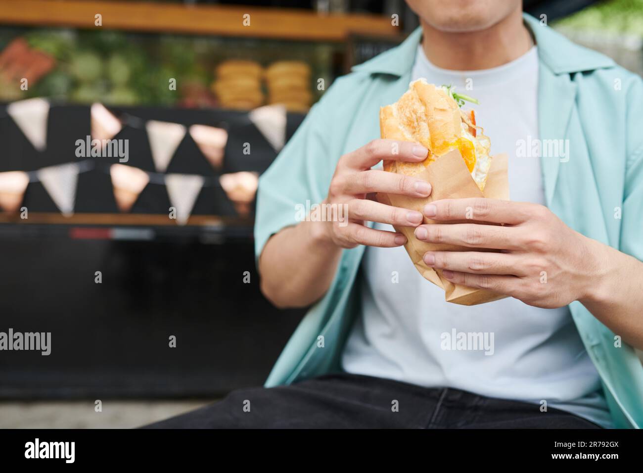 Nahaufnahme eines jungen hungrigen Mannes in Freizeitkleidung, der einen appetitlichen Hotdog in Papier hält, während er vor der Kamera gegen einen Street Food Truck steht Stockfoto