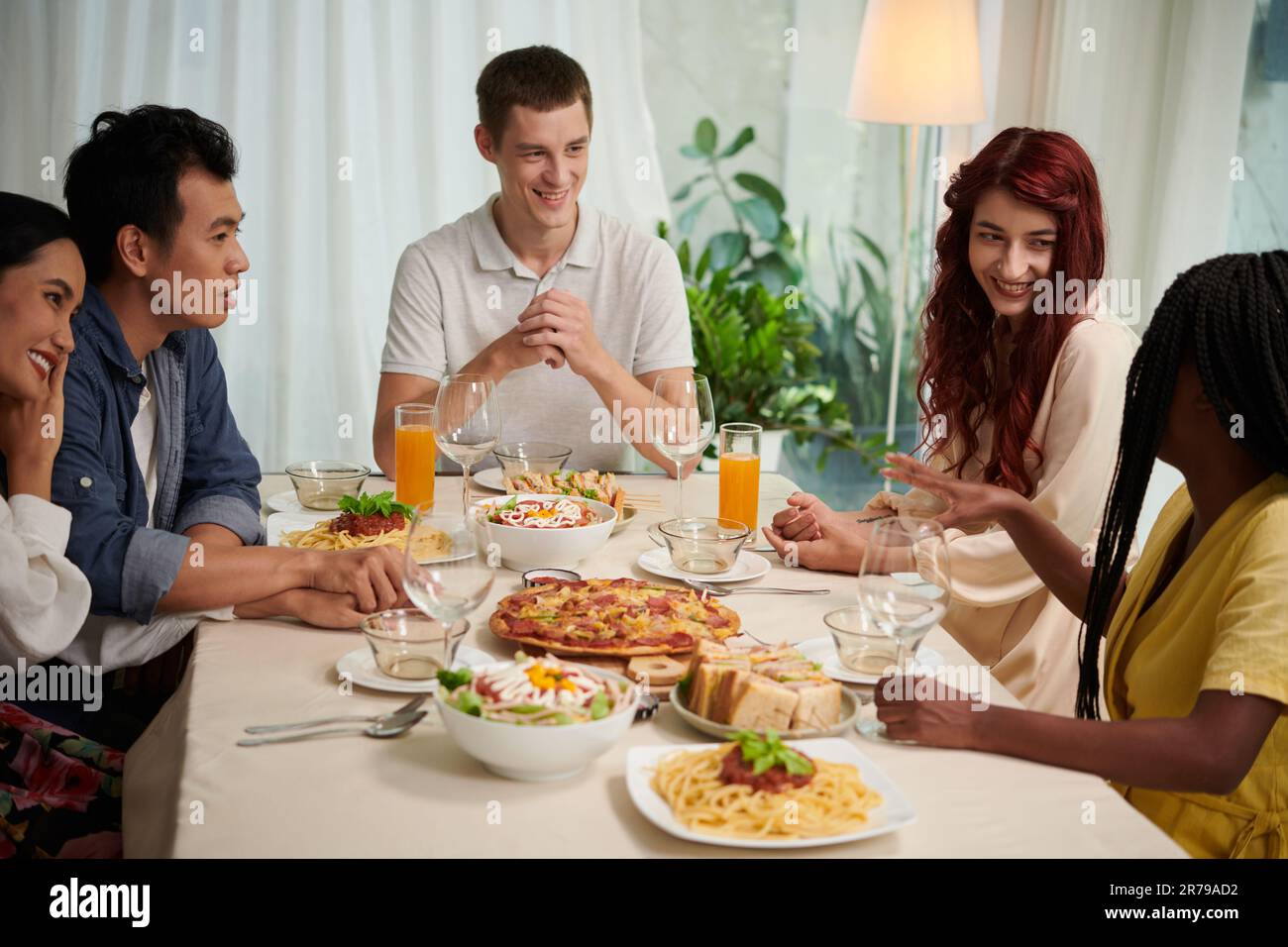 Eine Gruppe junger interkultureller Menschen in eleganter Freizeitkleidung, die sich am Serviertisch unterhalten und ein Abendessen mit hausgemachtem Essen genießen Stockfoto