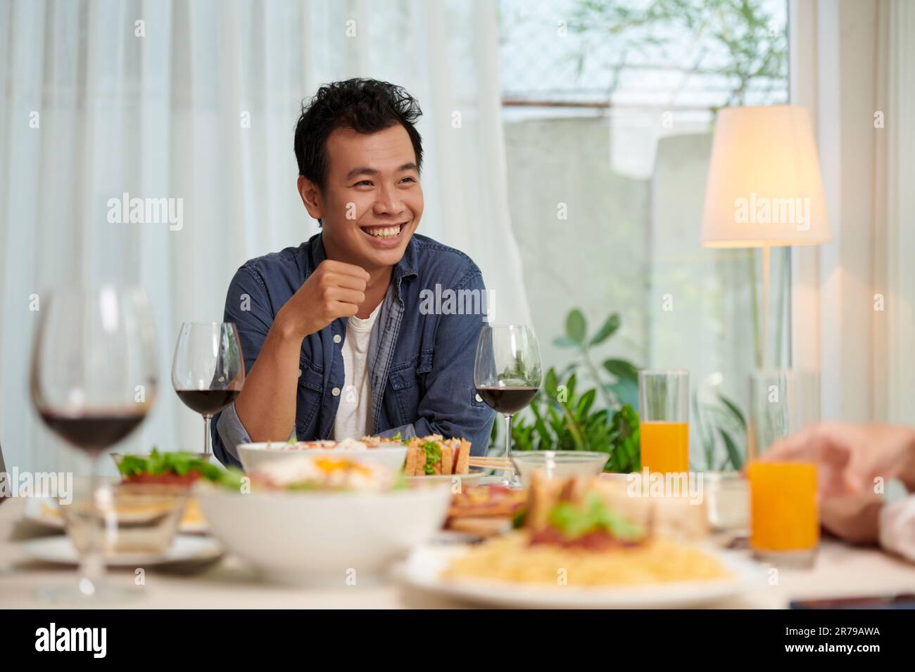 Junger, fröhlicher Asiate in Freizeitkleidung, der beim Abendessen am Serviertisch sitzt und seinen Freund während der Unterhaltung ansieht Stockfoto