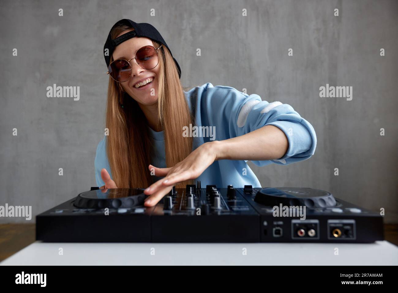 Ein junges, hübsches, langhaariges DJ-Mädchen in einem blauen Pullover, einer schwarzen Baseballkappe und einer Sonnenbrille posiert und mischt Musiktitel auf einer schwarzen DJ-Konsole. Studio Sho Stockfoto