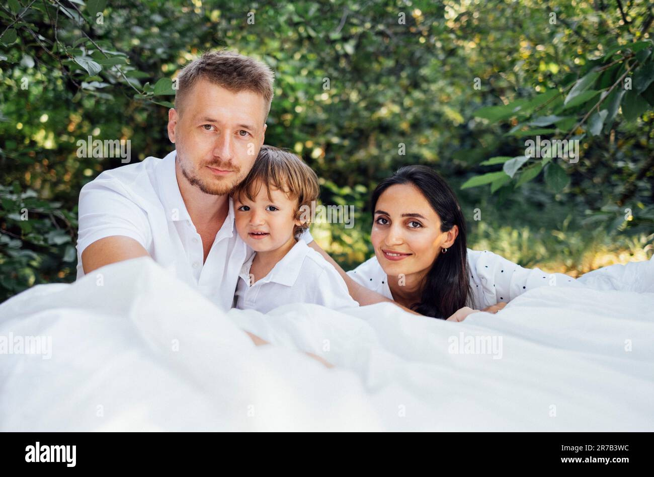 Porträt einer glücklichen, süßen Familie im Garten. Mom, Dad und Baby posieren und schauen in die Kamera. Lächelnde Eltern und ihr kleiner Sohn im Licht Stockfoto