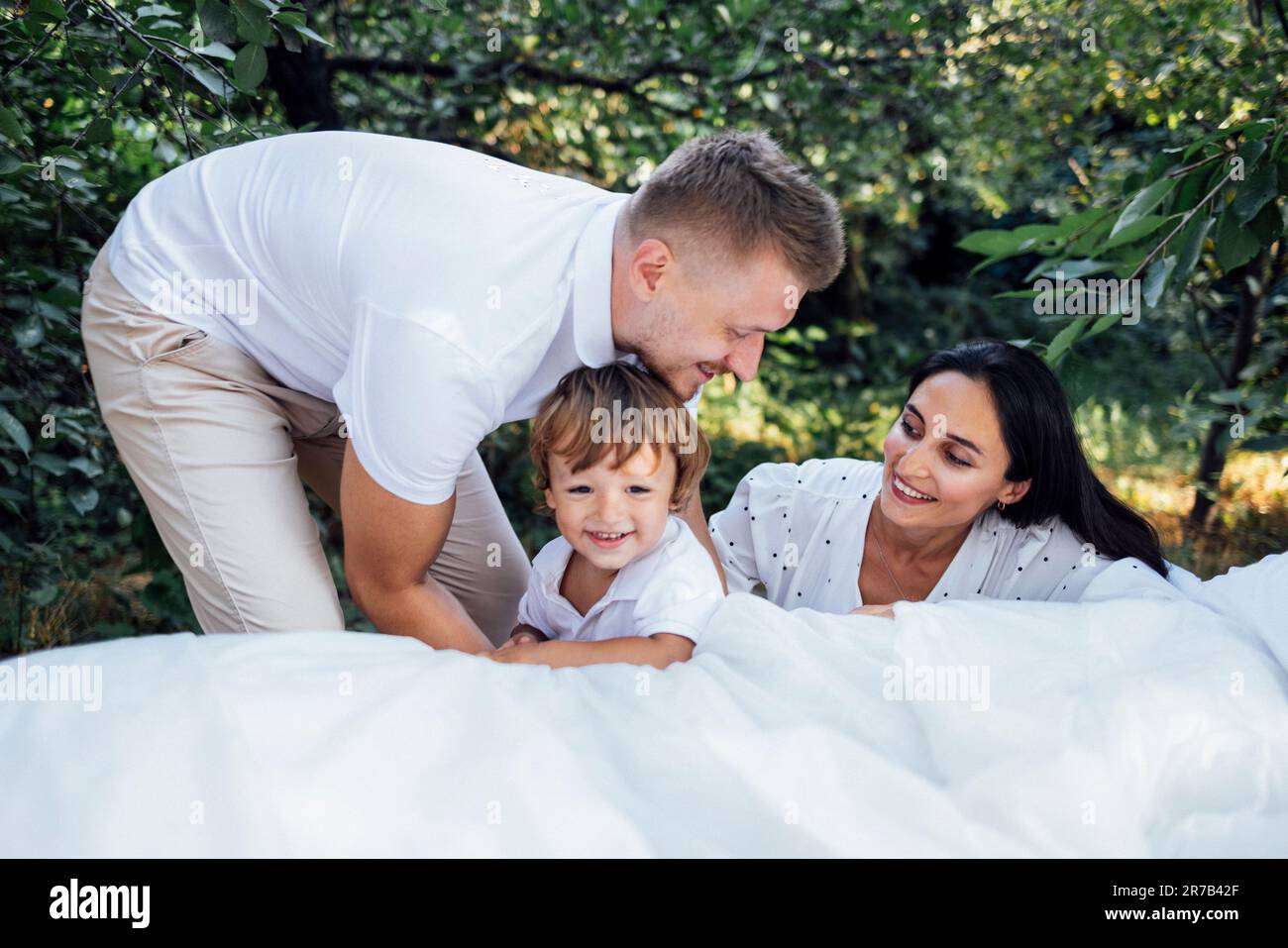 Porträt einer glücklichen, süßen Familie im Garten. Mom, Dad und Baby posieren und schauen in die Kamera. Lächelnde Eltern und ihr kleiner Sohn im Licht Stockfoto
