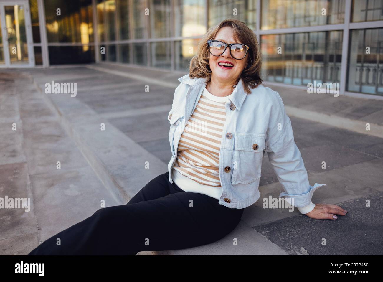 Eine Frau mittleren Alters mit Brille sitzt draußen auf der Treppe. Eine ältere Frau in lässiger Kleidung lacht und schaut in die Kamera. Terrassengebäude mit Stockfoto