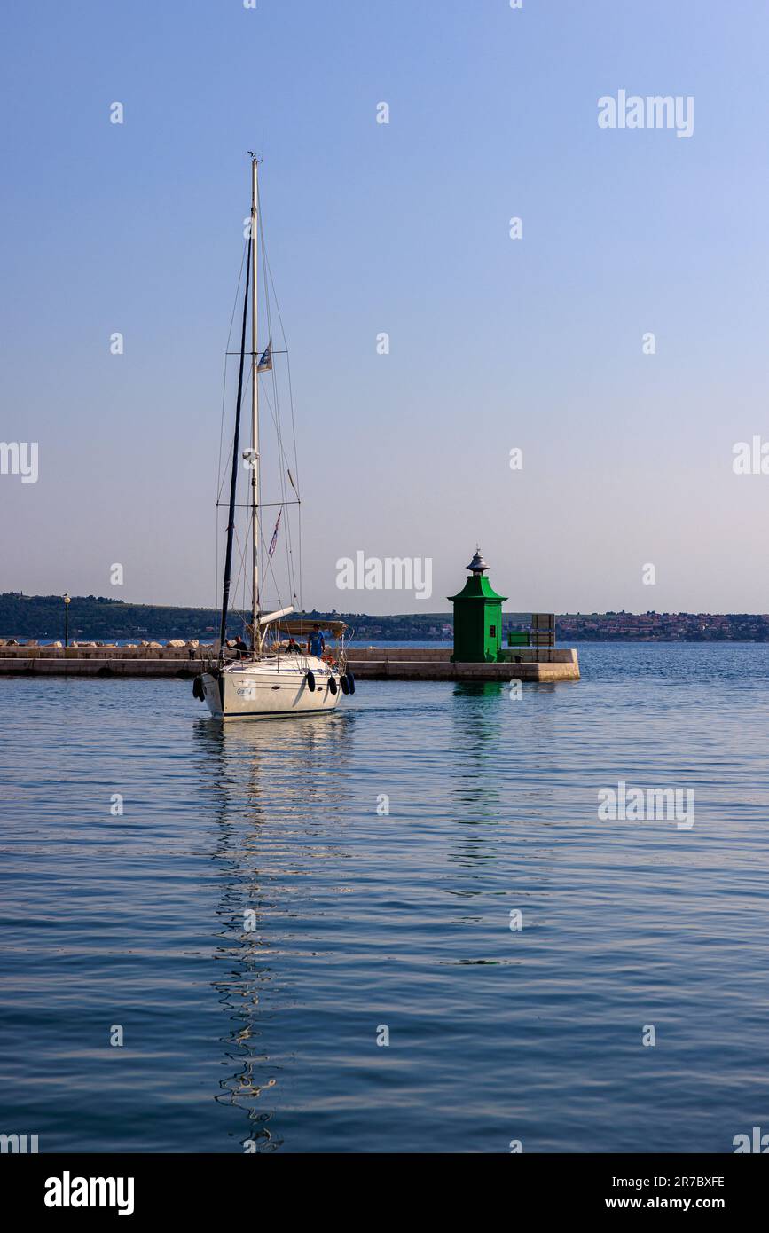 Eine weiße Yacht segelt in das ruhige Wasser des piran Hafens vorbei am grünen Hafen Leuchtturm, der den Eingang bewacht Stockfoto