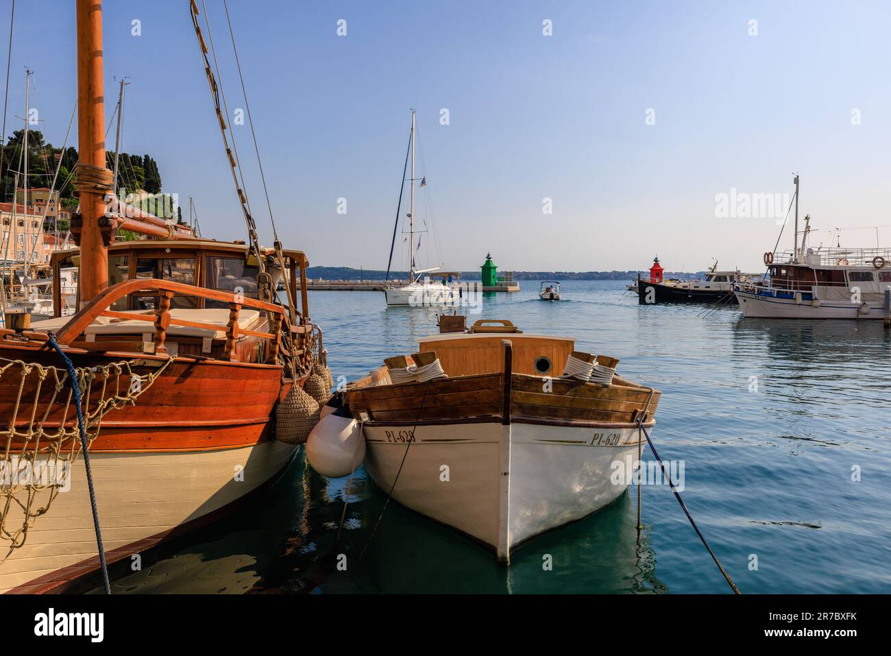 Hölzerne Boote, die auf einem ruhigen Meer im Hafen von Piran festgemacht sind, während eine Yacht an den grünen und roten Leuchttürmen des Hafens vorbeisegelt Stockfoto
