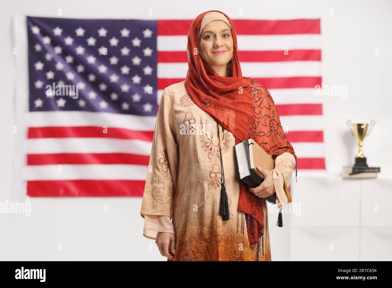 Das ganze Porträt einer arabischen Frau in einem Hidschab, die vor der amerikanischen Flagge steht und Bücher hält Stockfoto