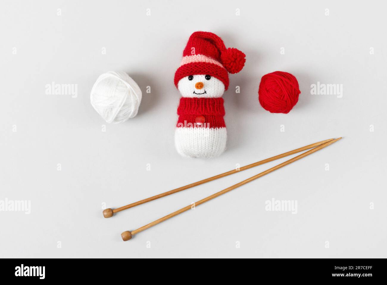 Gestrickter Schneemann mit rotem Hut und Pullover mit Fäden und Stricknadeln auf grauem Hintergrund. Babysachen und Accessoires. Geschenk für den Winter Stockfoto