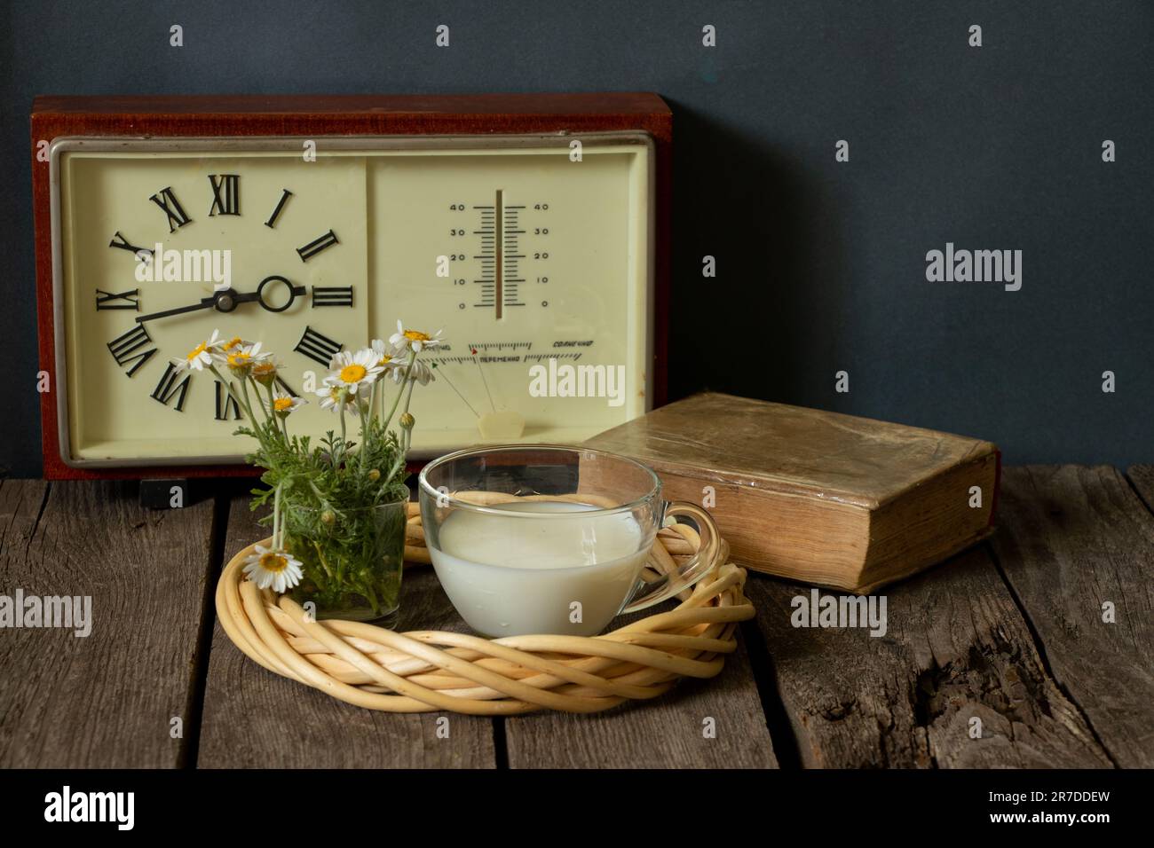 eine tasse Milch und eine Vase mit Gänseblümchen in der Nähe der Uhr mit einem Buch auf einem Holztisch Stockfoto