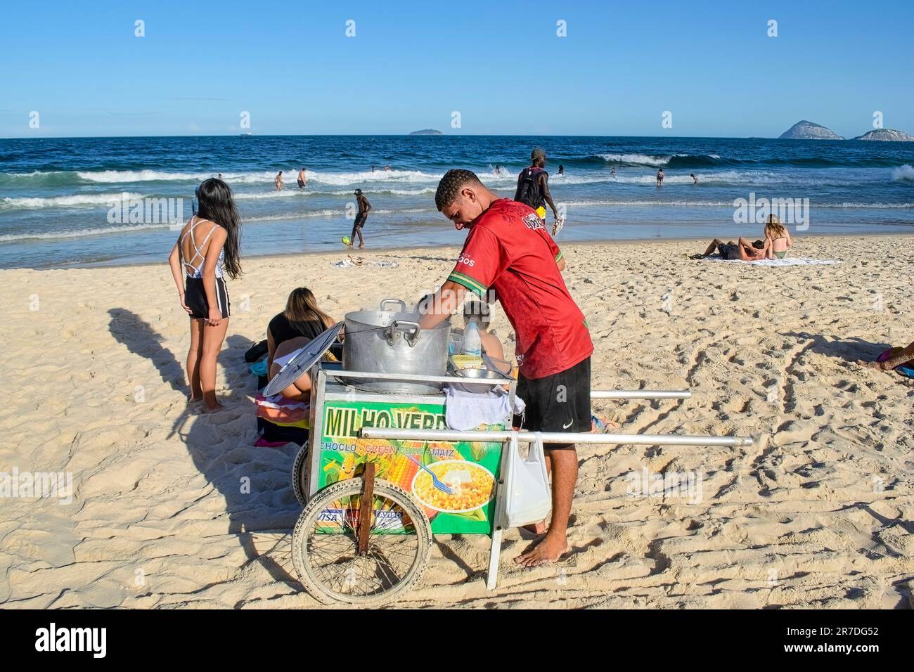 Rio de Janeiro, Brasilien - 25. Mai 2023: Ein Mann steht hinter einem Karren mit der Aufschrift Milho Verd am Copacabana Beach, mit einem Topf auf dem Karren. Strandbesucher und Stockfoto