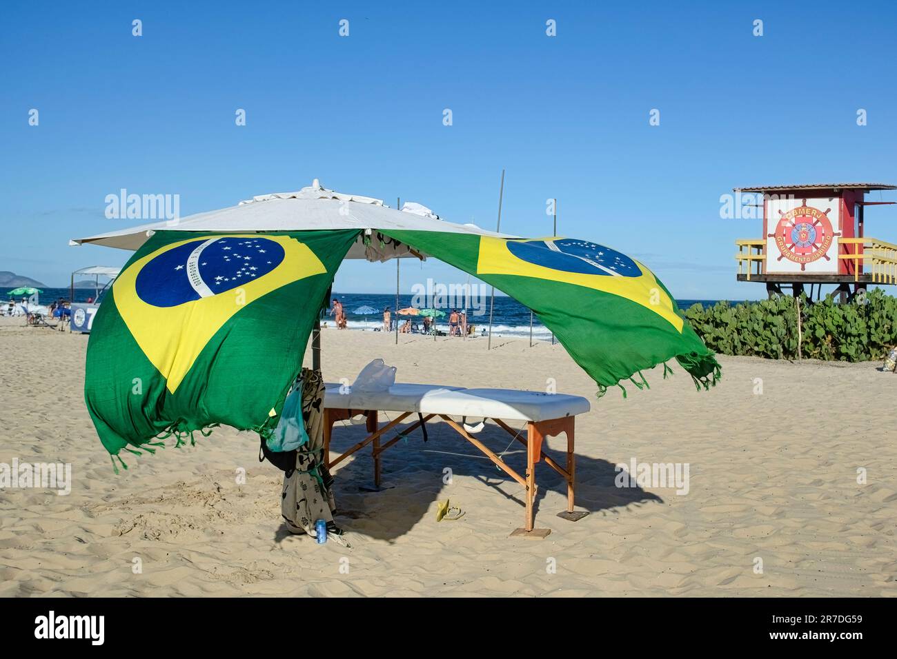 Rio de Janeiro, Brasilien - 25. Mai 2023: Ein Sonnenschirm mit brasilianischen Flaggen steht auf dem Sand und bietet Schatten für eine Bank. Die Leute entspannen sich an den s Stockfoto
