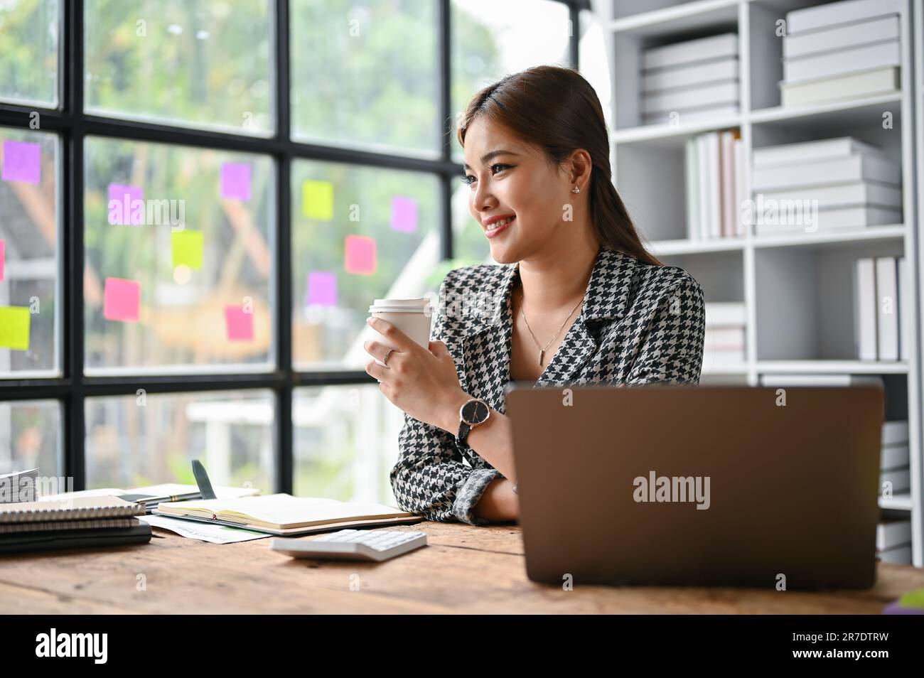Eine attraktive asiatische Geschäftsfrau der Millennials sitzt in einem trendigen Anzug an ihrem Schreibtisch und träumt, während sie ihren Morgenkaffee trinkt. Stockfoto