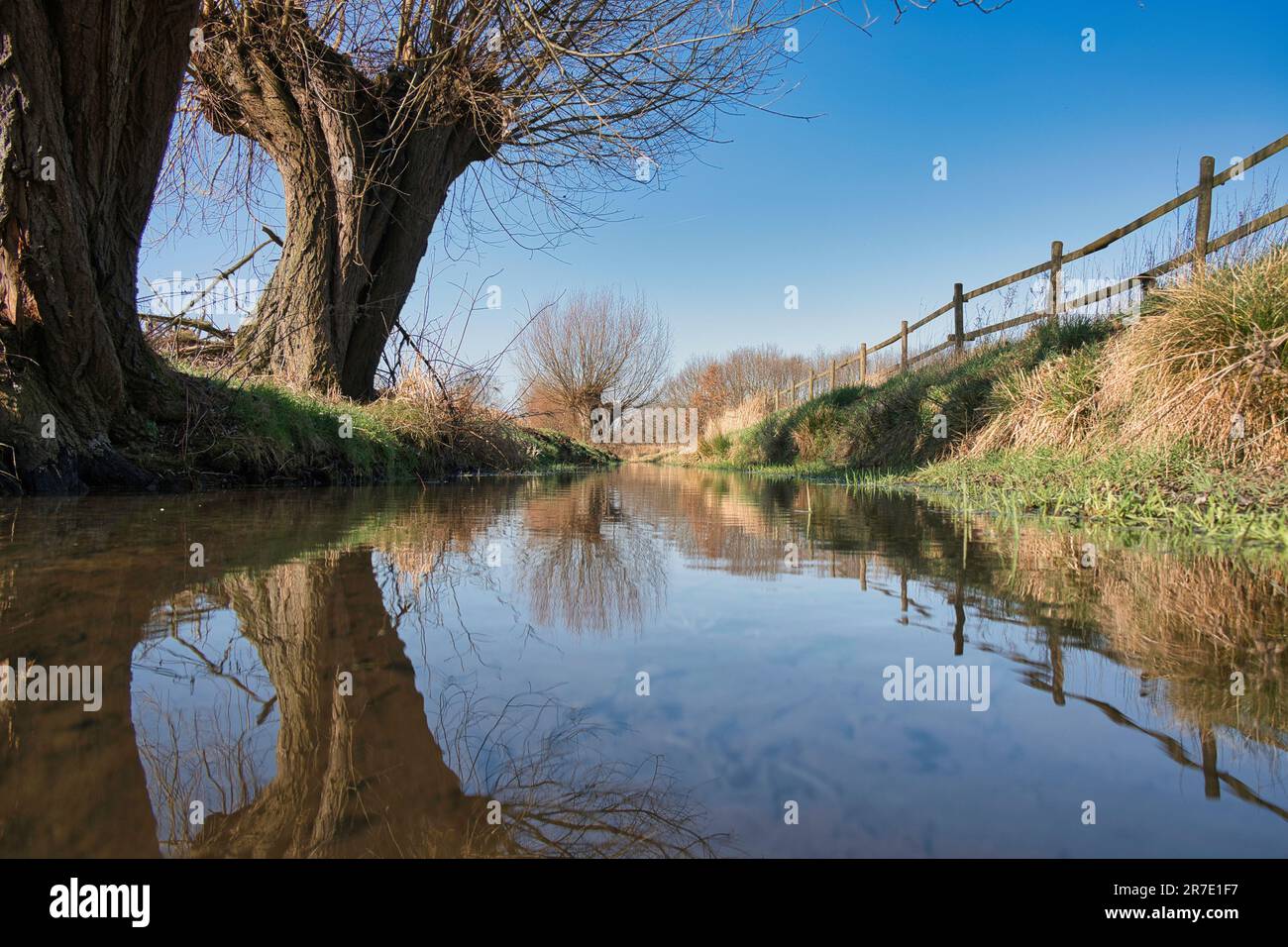 Eine ruhige Landschaft eines Flusses auf dem Land, mit einem Zaun auf einer Seite und Bäumen, die sich im stillen Wasser spiegeln Stockfoto