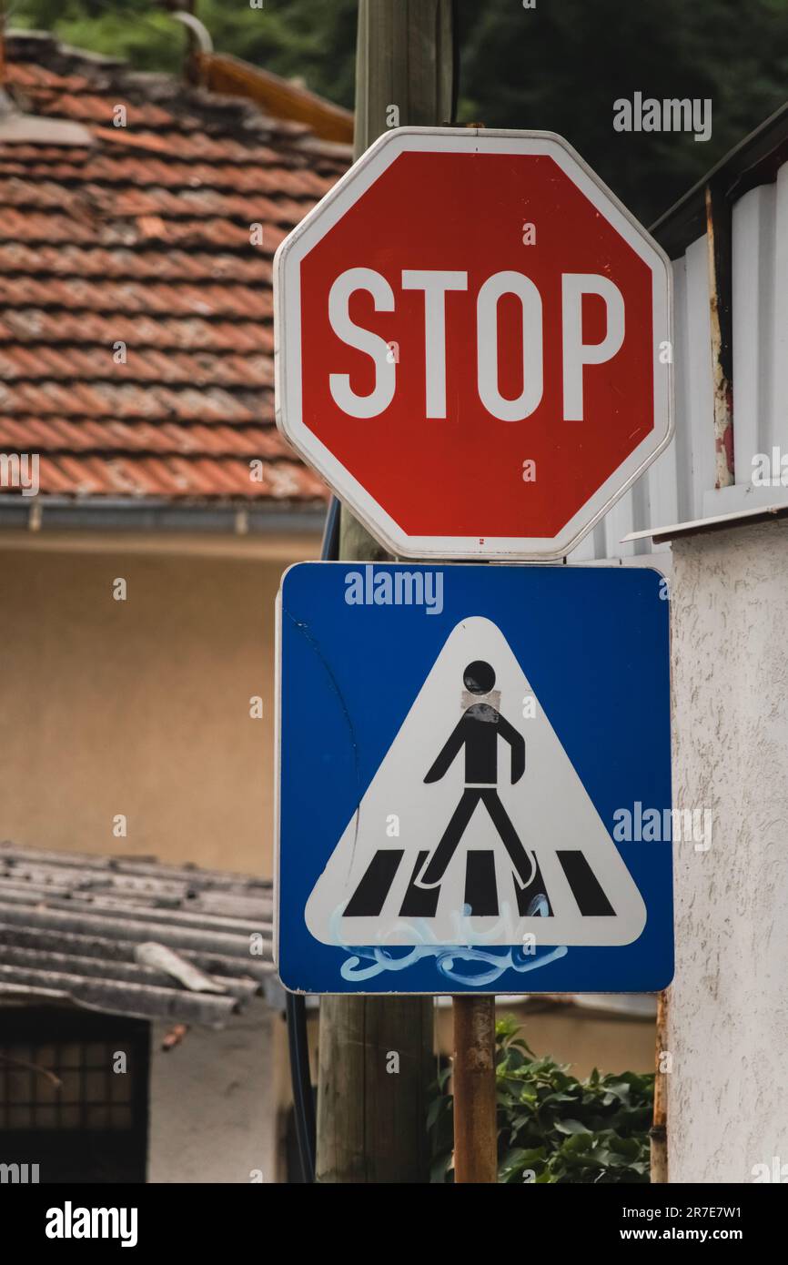 Zwei Metallschilder für Stopp und Überquerung in Rot, Blau und Schwarz zwischen Häusern mit Bäumen im Hintergrund Stockfoto