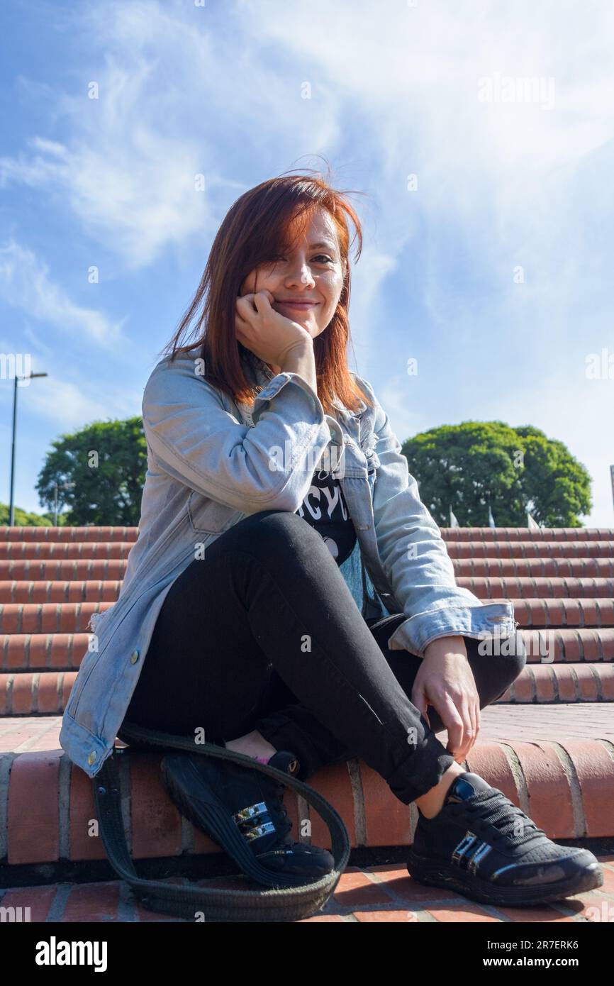 Junge lateinerin kolumbianischer Herkunft mit roten Haaren, die in die Kamera schaut, im Park sitzt, in die Kamera schaut und ihre Beine überkreuzt, sie Stockfoto