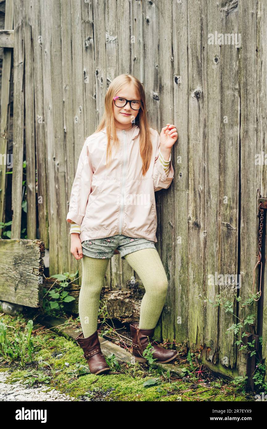 Outdoor-Modeporträt eines hübschen kleinen Mädchens, das eine Brille, einen pinkfarbenen regenmantel, grüne Strumpfhosen und braune Lederstiefel trägt Stockfoto
