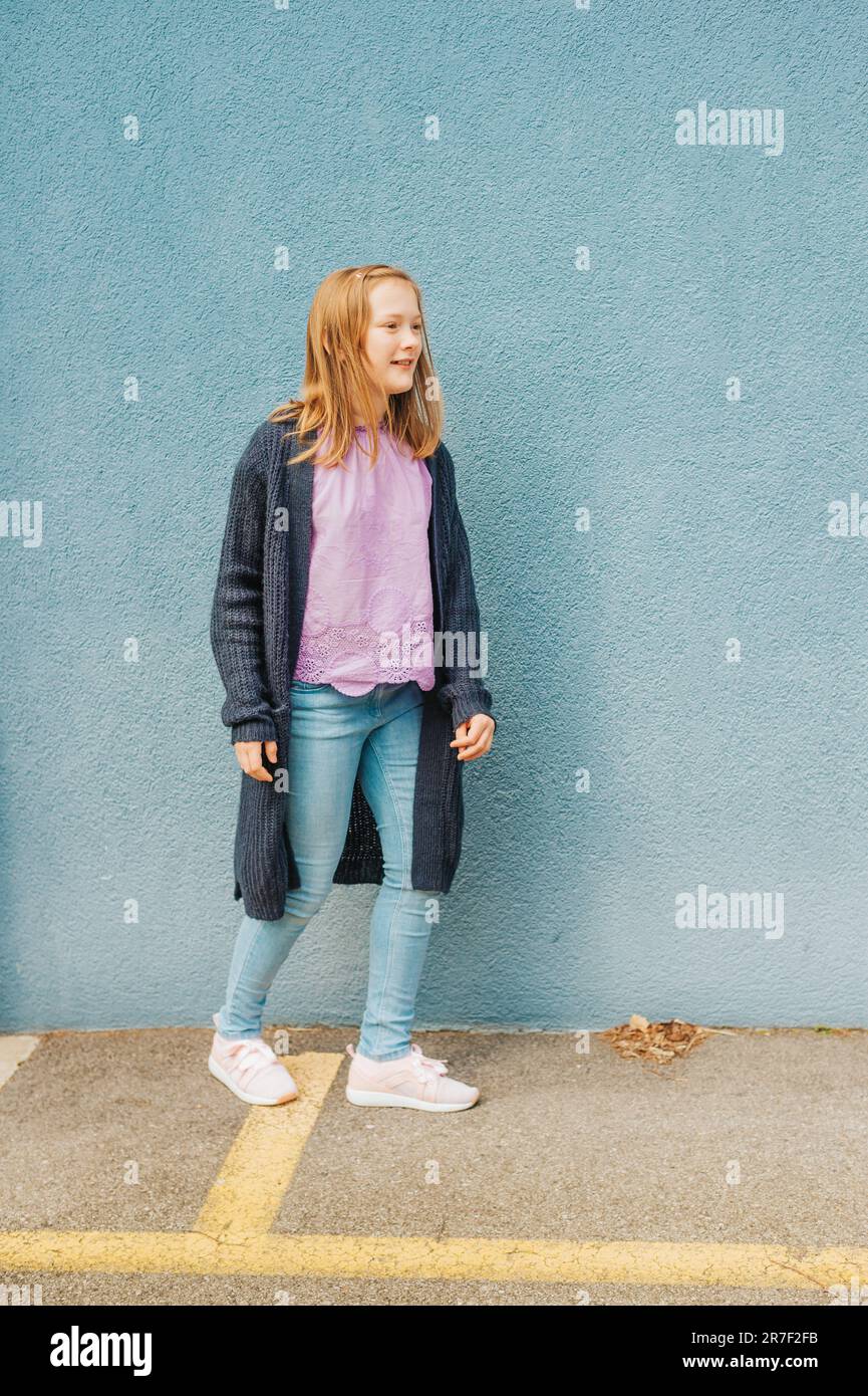 Außenporträt eines hübschen jungen 10-jährigen Mädchens, das eine gestrickte lange Jacke, Jeans und pinke Turnschuhe trägt und neben blauem Hintergrund posiert Stockfoto