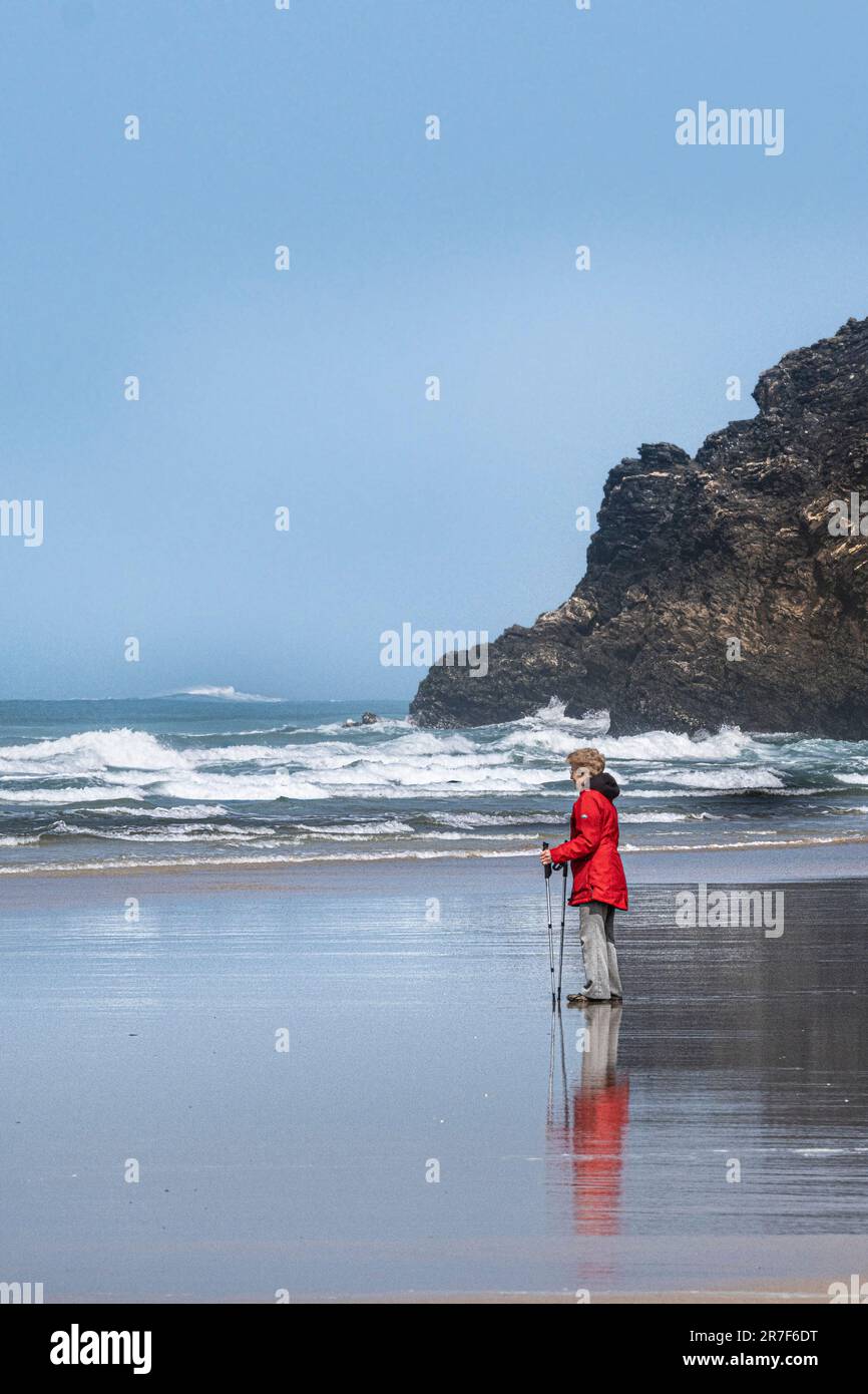 Eine reife Frau, die einen leuchtend roten Mantel trägt und Wanderstöcke am Ufer des Mawgan Porth Beach in Cornwall im Vereinigten Königreich verwendet. Stockfoto