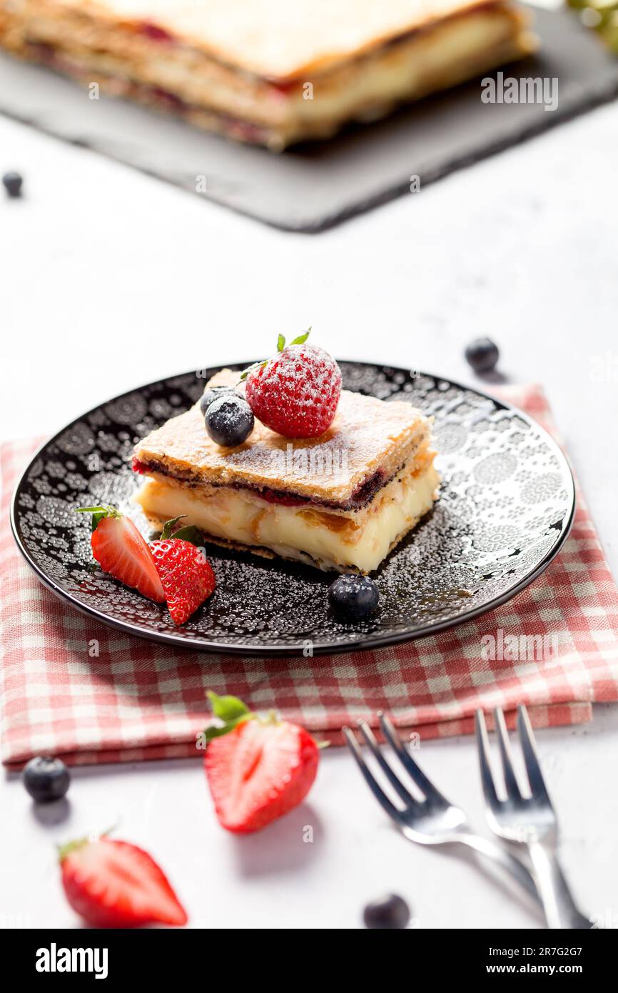 Ein Stück Napoleonkuchen mit Mohnsamen und Kirschen, dekoriert mit Erdbeeren und Blaubeeren auf hellem Hintergrund. Schließen Stockfoto