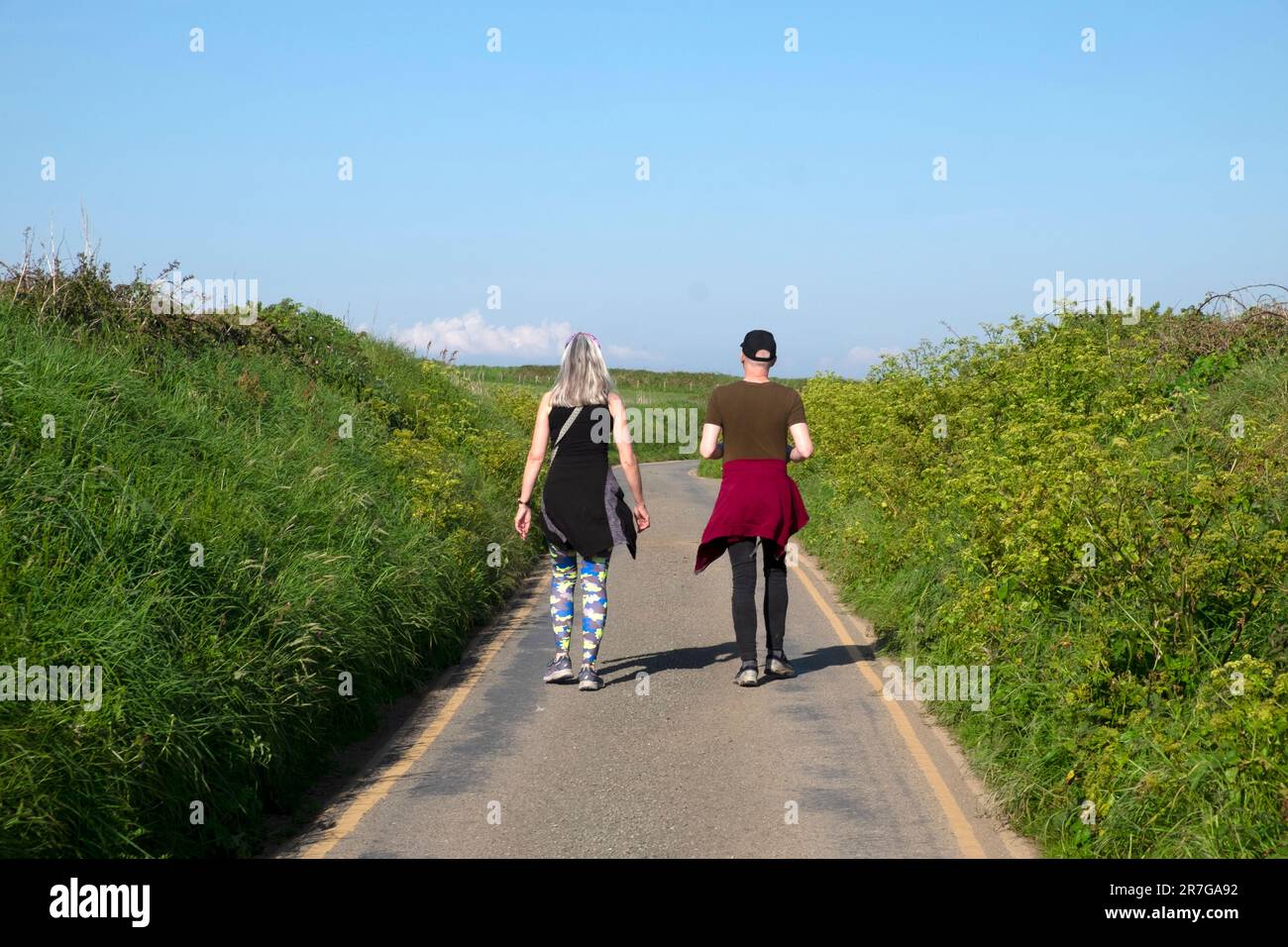 Rückansicht von zwei Freunden, die im Sommer an einem sonnigen Tag im Frühjahr 2023 auf einer Landstraße spazieren. Pembrokeshire Wales UK KATHY DEWITT Stockfoto