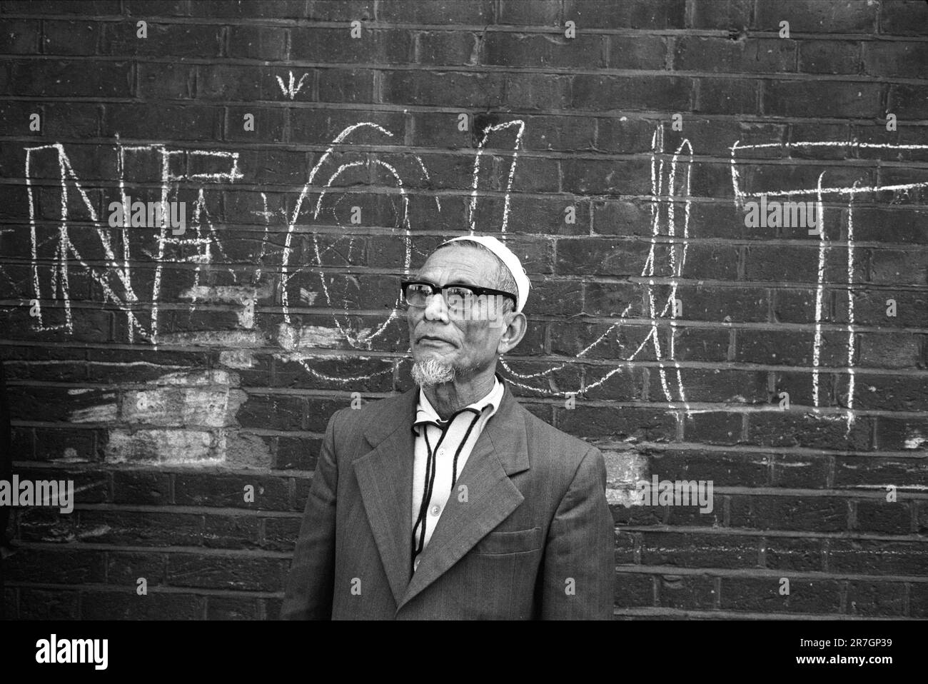 Ein älterer asiatischer Mann, ein Einwanderer in der Gegend, beobachtet das Sonntagmorgen-Leben in BricLane. Das Graffiti "NF OUT" bezieht sich auf die Front National, die versuchte, Unterstützung für ihre Einwanderungspolitik zu gewinnen. Whitechapel, East London, England, ca. 1978. UK 1970S HOMER SYKES Stockfoto