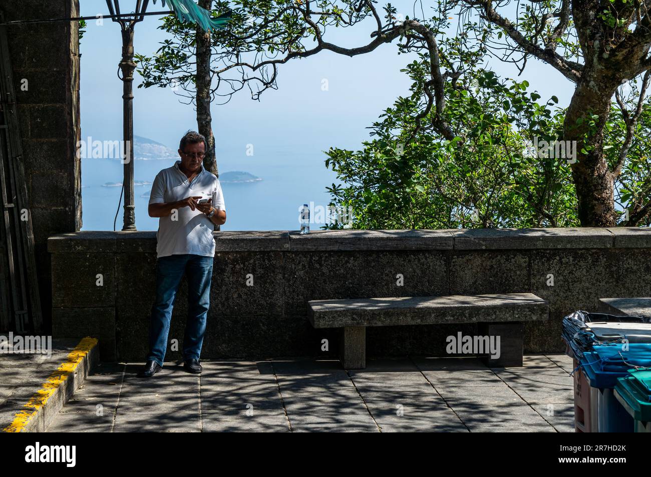 Ein Tourist nimmt sich einen Moment Zeit, um an einem sonnigen Sommertag sein Smartphone in der Nähe einer kurzen Mauerbeobachtungsplattform in der Nähe des Gipfels des Corcovado zu überprüfen. Stockfoto