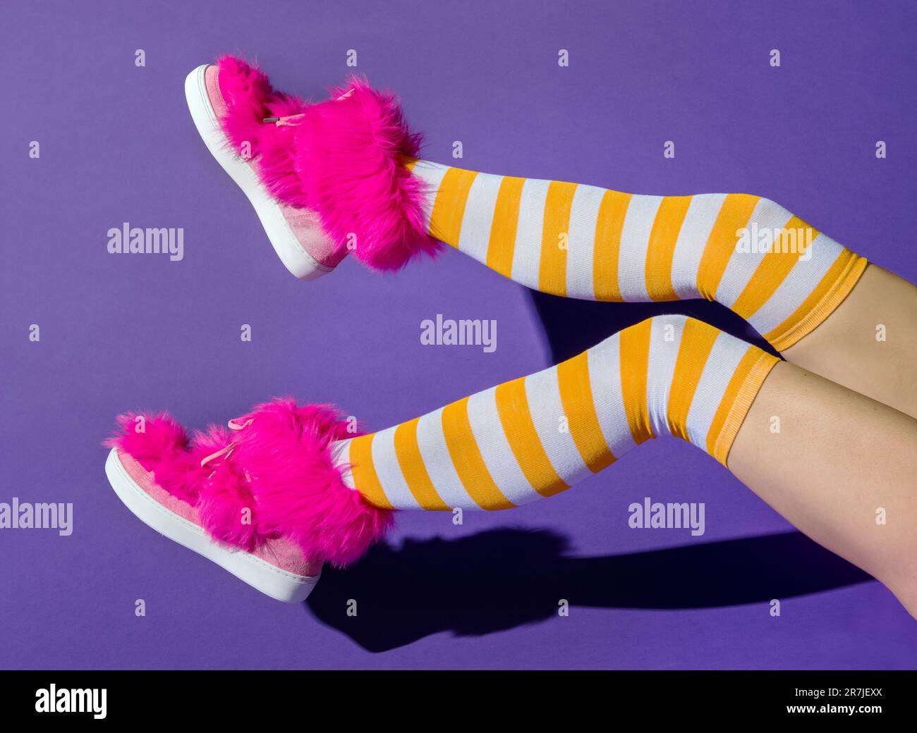 Seitenansicht einer anonymen Frau mit einem Schatten aus hochgezogenen Beinen in gelben langen Socken und pinkfarbenen Fuchsieschuhen auf violettem Hintergrund Stockfoto