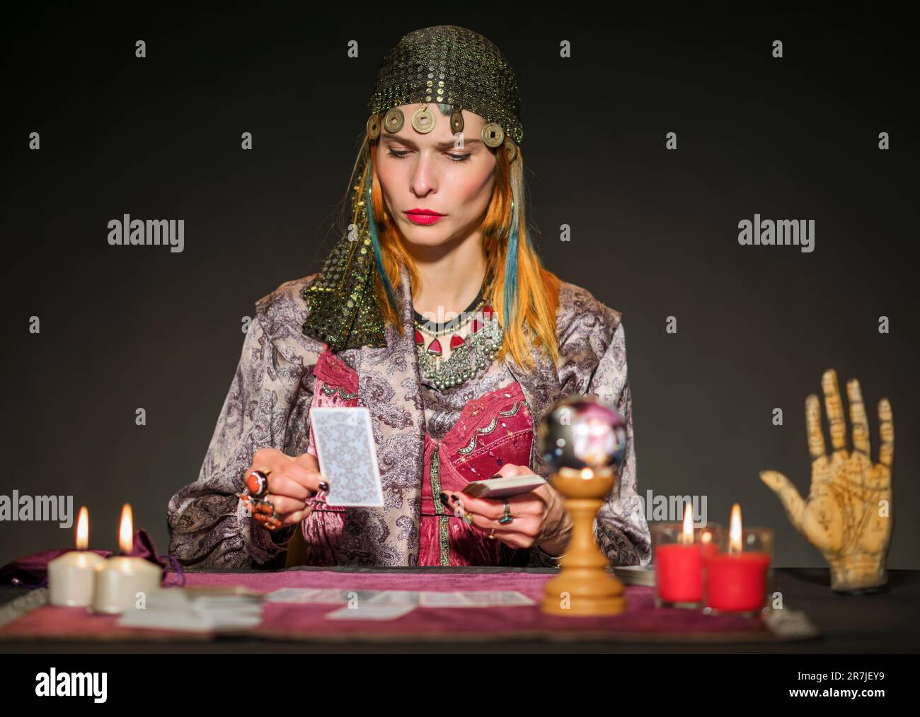 Fokussierter Wahrsager in authentischem Outfit und Münzkopfstück, der mit brennenden Kerzen am Tisch sitzt und mit Tarotkarten die Zukunft erraten kann Stockfoto