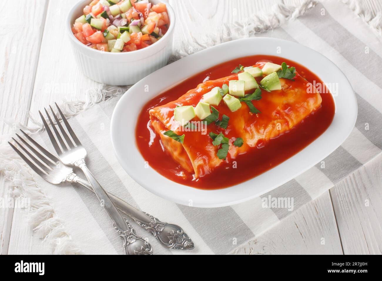 Leckere, nasse Burritos mit Rindfleisch, Reis und gekühlten Bohnen, verpackt in einer Tortilla, erstickt mit roter Sauce, auf dem Teller auf dem Holztisch. Stockfoto