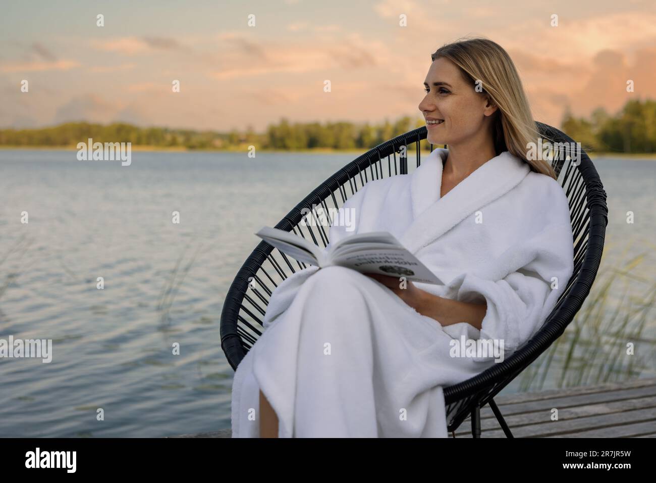 Frau im weißen Bademantel liest ein Buch im Stuhl auf der Fußgängerbrücke des Sees und genießt einen warmen Sommerabend Stockfoto