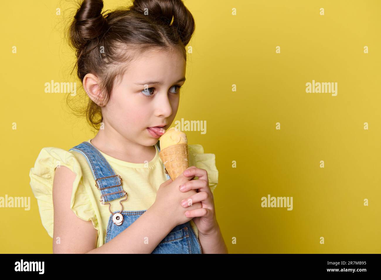 Nahaufnahme eines kleinen Mädchens, das eine Eiskugel in Waffeleisen leckt und einen Kopierbereich auf gelbem Hintergrund zur Seite blickt Stockfoto