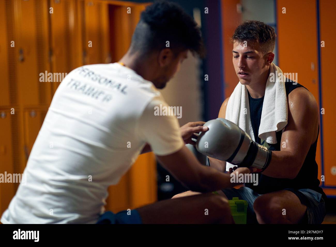 Junger Boxspieler mit Trainer im Umkleideraum des Fitnessstudios, der auf einer Bank sitzt. Der Personal Trainer bereitet den Spieler auf das Spiel vor. Stockfoto