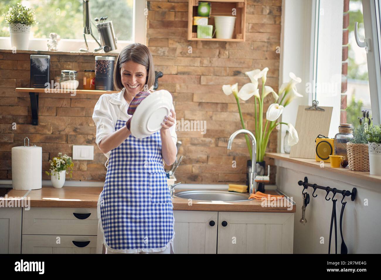 Eine strahlende Frau füllt den Raum mit ihrem strahlenden Lächeln, während sie fleißig sauberes Geschirr nach einem zufriedenstellenden Waschgang abwischt. Stockfoto