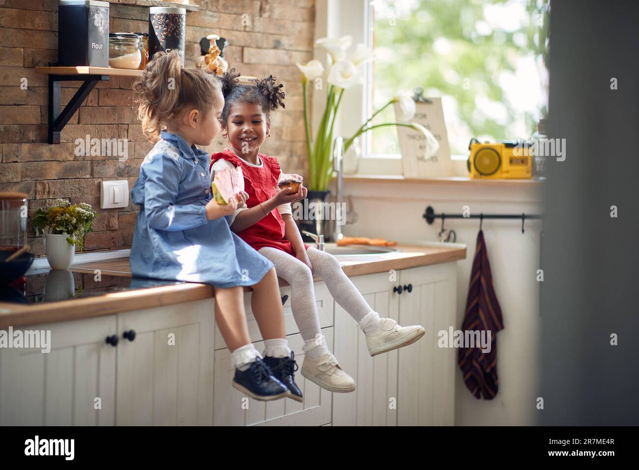 Zwei niedliche kleine Mädchen sitzen auf der Küchentheke, genießen einen Snack zusammen und essen eine Wassermelone und einen Muffin. Zuhause, Lebensstil, Familienkonzept Stockfoto