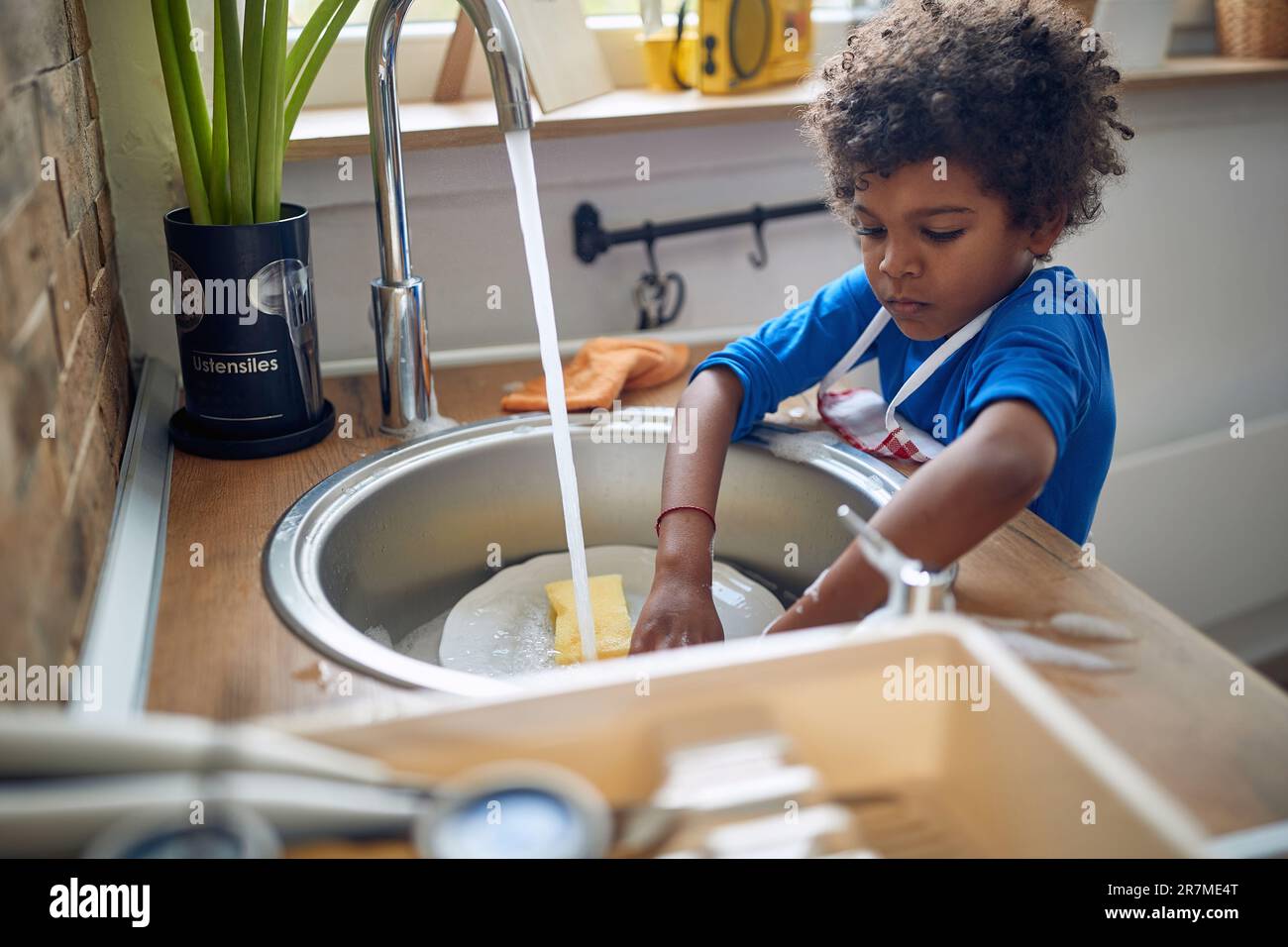 Der junge afroamerikanische Junge übernimmt die Verantwortung, mit großer Begeisterung Geschirr zu spülen. Mit einer bunten Schürze steht er selbstbewusst auf der si Stockfoto