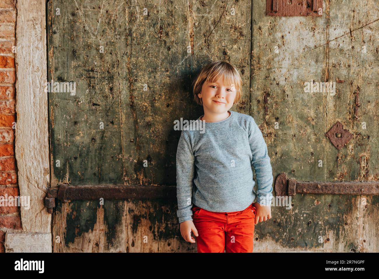 Außenporträt eines gutaussehenden kleinen Jungen mit roter Chino und hellblauem Pullover, stilvolles Kind, das gegen eine alte alte alte Wand posiert. Mode für kleines Chi Stockfoto